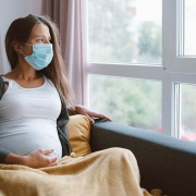 Pregnant woman sitting on a couch, looking out the window with a concerned expression, wearing a mask.