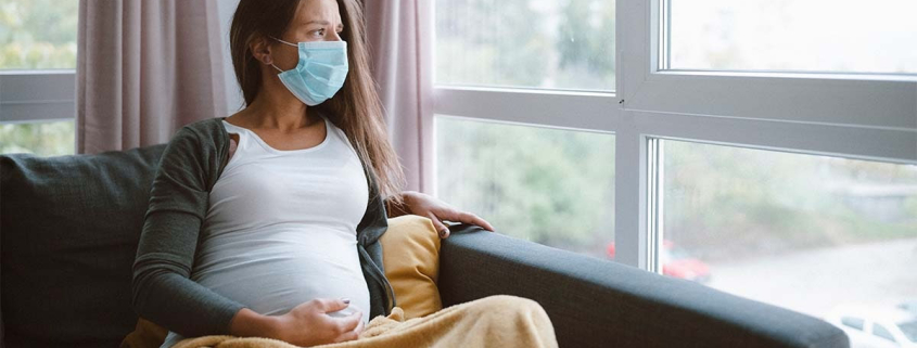 Pregnant woman sitting on a couch, looking out the window with a concerned expression, wearing a mask.